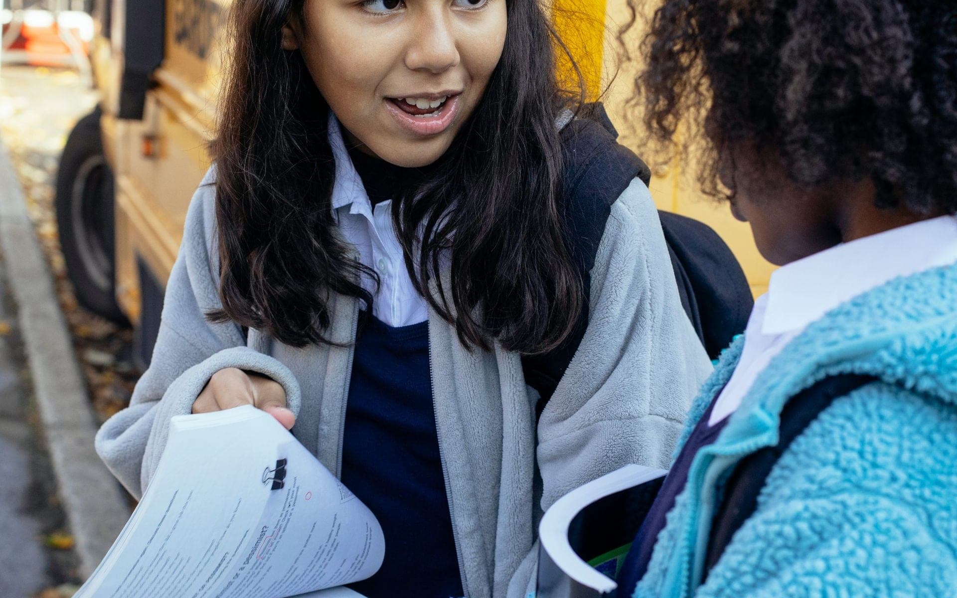 Two girls with backpacks talking outside a yellow school bus